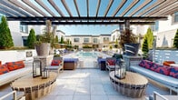 This expansive outdoor area showcases a stunning aquatic complex with multiple pools, creating a resort-like atmosphere in the heart of an urban residential development. The main pool, rectangular in shape, is flanked by a smaller, more intimate pool area, offering variety for swimmers. The poolside is adorned with an array of comfortable seating options, including numerous blue lounge chairs and white in-pool loungers, perfect for relaxation and sunbathing.

The architectural design of the surrounding buildings is modern and sleek, featuring white brick facades, large windows, and flat rooflines. Wooden pergolas add a touch of warmth and provide partial shade to certain areas. The landscaping is meticulously maintained, with evergreen trees, potted plants, and climbing vines softening the hardscaping and adding natural beauty to the space.

The pool deck is constructed of light-colored pavers, creating a clean and spacious feel. Safety features are evident, with pool rules clearly displayed and depth markers visible. The overall ambiance is one of luxury and tranquility, offering residents a private retreat for relaxation and socialization. This thoughtfully designed space seamlessly blends functionality with aesthetic appeal, making it a standout amenity for the residential complex.
