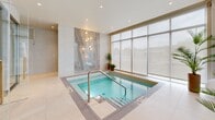 Located at 3000 Rue Modugno in Montréal, this sophisticated spa facility offers a refined wellness experience within a thoughtfully designed single-level space. The interior is defined by a clean, contemporary aesthetic, featuring large floor-to-ceiling windows that flood the area with natural light and offer views of the surrounding outdoors, creating a calm and serene atmosphere.

At the heart of the spa is a pristine indoor pool with a tiled surround and stainless steel entry rails, ideal for hydrotherapy. Adjacent to the pool, a separate hot tub is framed by an elegant marble accent wall adorned with soft globe lighting, adding a luxurious focal point to the space.

The spa also includes a wood-lined sauna enclosed in frameless glass panels with gold-toned hardware, providing a warm, traditional Finnish sauna experience. Nearby, a glass-enclosed steam room offers an additional therapeutic option, both accessible directly from the main pool area.

Throughout the space, large-format cream-toned floor tiles, recessed lighting, and glass partitions contribute to an open, uncluttered design. Potted tropical plants are thoughtfully placed to introduce organic warmth to the otherwise minimalist palette. The overall ambiance is one of understated luxury — clean, bright, and purpose-built for relaxation and wellness.