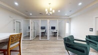 Located at 655 W Mallard Creek Church Rd in Charlotte, NC, this single-floor professional office space combines modern design with practical functionality. The interior is finished with light wood-look flooring and tray ceilings throughout, creating a clean, polished aesthetic.

A standout feature is the private office pod area, where multiple glass-enclosed individual workstations are arranged side by side. Each pod is outfitted with a dedicated desk, monitor, and ergonomic chair, offering a quiet, focused environment for individual work. The glass walls and white framing provide visual openness while maintaining privacy.

The lounge areas are furnished with rich emerald green velvet armchairs arranged around low coffee tables atop neutral area rugs. Framed artwork on the walls and soft recessed lighting contribute to a welcoming, professional atmosphere. One lounge area includes a stylish chandelier accent overhead.

A breakroom or conference area features a large white high-top table surrounded by carved-back bar stools, a wall-mounted flat-screen television, and an undercounter appliance, making it suitable for team gatherings or casual meetings.

Throughout the space, tray ceilings, in-ceiling speakers, and recessed lighting are consistent architectural details that reinforce the space's refined, professional character. Restroom facilities are also accessible on this floor.