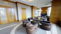 This single-floor space presents a well-appointed lounge or lobby area designed with a refined, contemporary aesthetic. The room is anchored by a carefully curated seating arrangement, including a striped sofa accented with yellow throw pillows and several rounded, upholstered barrel chairs in muted purple-brown tones, all centered around a sleek, dark circular coffee table. The flooring features a patterned area rug with sweeping curved lines, adding visual interest and defining the seating zone.

Warm honey-toned wood paneling frames key architectural elements throughout the space, including pilasters and door surrounds, providing a rich contrast against the light grey walls. A fireplace with a dark surround serves as a focal point on one wall, flanked by subtle wall-mounted sconces that contribute to the ambient lighting. Natural light enters through windows visible toward the rear of the space, complementing the recessed ceiling lighting and a softly illuminated tray ceiling detail above.

A framed artwork piece decorates the wall near the fireplace, adding a curated touch to the overall design. The space conveys a professional yet welcoming atmosphere suited for communal or hospitality use.