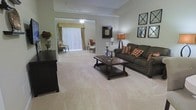 Located at 20 Bur Reed Road in Delaware, Ohio, this single-floor residence offers a well-appointed and comfortable living environment designed with both functionality and style in mind. The open layout creates a sense of generous space throughout, with rooms flowing naturally into one another for easy everyday living. The main living area is characterized by plush, light-colored wall-to-wall carpeting that adds warmth underfoot, complemented by neutral wall tones that create a calm and inviting ambiance. The living room is thoughtfully proportioned, offering ample space for seating arrangements and entertaining. Crown molding details along the upper walls add a refined architectural touch. A wide sliding glass door framed by vertical blinds and a valance connects the interior to a sunlit outdoor patio area, allowing natural light to fill the space and providing a seamless indoor-outdoor connection. The adjacent area, visible through an open passageway, extends the living space further and features access to the same outdoor patio, enhancing the overall sense of openness. Decorative wall art and warm accent lighting contribute to the cozy, welcoming character of the home, making it a comfortable and practical single-level living space in Delaware, Ohio.