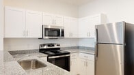 Located at 1401 Davenport Blvd in Davenport, Florida, this single-story, 4-bedroom, 2-bathroom home offers a well-designed layout with clean, modern finishes throughout. The kitchen serves as a central highlight, featuring crisp white shaker-style cabinetry, granite countertops, a peninsula with a built-in sink, and a full suite of stainless steel appliances including a refrigerator, electric range, microwave, and dishwasher. Recessed lighting and an open sightline to the living area enhance the bright, airy feel of the space. Light wood-style flooring runs throughout the home, contributing to a cohesive and contemporary aesthetic. The bedrooms are generously proportioned, with the primary bedroom offering ample space and natural light through a large window. Three closets provide practical storage, including a walk-in closet with wire shelving. Both bathrooms are finished with granite-topped white vanities and tiled walk-in showers. The overall palette of soft whites and light neutrals creates a clean, welcoming ambiance. The home's straightforward single-floor plan ensures easy accessibility and a comfortable flow between all rooms.
