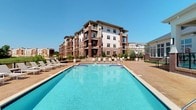 This residential community showcases a striking multi-story building constructed with classic brick and neutral stucco exteriors, offering a timeless architectural aesthetic. The standout feature of the outdoor amenity area is a generously sized swimming pool with crystal-clear blue water, ideal for recreation and relaxation. The surrounding pool deck is finished with a warm-toned paved surface and is well-equipped with multiple lounge chairs and seating areas, providing ample space for residents to unwind under open skies. Mature landscaping, including neatly trimmed shrubs and planters, lines the perimeter of the deck, adding greenery and a welcoming atmosphere to the outdoor space. The building itself features private balconies on upper floors, allowing residents to enjoy outdoor views from their own space. Large windows throughout the facade ensure generous natural light reaches interior units. The overall ambiance of the community is clean, modern, and well-maintained, blending classic brick detailing with contemporary design elements to create an inviting environment suited to comfortable residential living.