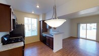 This single-floor space showcases an open-concept layout that integrates the kitchen and living area into one cohesive, functional environment. The kitchen is appointed with rich, dark espresso cabinetry that provides ample storage, complemented by granite-style countertops that extend to a peninsula with a built-in sink. The peninsula serves as a natural divider between the kitchen and living space while also offering the potential for casual seating or additional workspace. A stylish brushed-nickel pendant light hangs above the peninsula, adding a contemporary focal point to the space. Black appliances, including a refrigerator and dishwasher, are seamlessly integrated into the cabinetry. Warm hardwood flooring extends throughout the open area, contributing to a cohesive and inviting atmosphere. Recessed lighting in the kitchen provides functional illumination, while a window fitted with horizontal blinds allows natural light to filter into the space, brightening the neutral-toned walls. The living area beyond the kitchen offers an open, versatile space that benefits from the natural light and the warm tones established by the flooring and cabinetry.