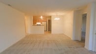 This single-floor space is designed with an open-concept layout that seamlessly connects the main living area, dining space, and kitchen. The living area is finished with light-toned wall-to-wall carpeting, creating a warm and neutral foundation that extends throughout the space.

The kitchen is positioned toward the rear and features a peninsula counter that serves as a natural divider between the cooking and living areas. Warm honey-toned wood cabinetry lines the kitchen walls, complemented by a white range and a stainless steel refrigerator. The kitchen benefits from its own ceiling light fixture, ensuring the workspace remains well-lit.

Adjacent to the kitchen, the dining area is distinguished by a decorative chandelier that adds a subtle touch of elegance to the open space. A doorway visible from this area leads to an additional room, providing functional separation within the otherwise open floor plan.

The walls throughout are finished in a clean, neutral off-white tone, enhancing the sense of openness. The overall ambiance is calm and understated, offering a versatile blank canvas well-suited for a variety of interior arrangements.