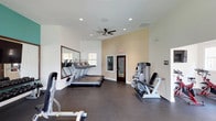 This fitness studio is a thoughtfully designed, single-floor facility that offers a wide range of exercise equipment to accommodate varied workout routines. The space is characterized by its clean, modern aesthetic, featuring crisp white walls accented by warm beige and bold teal feature walls that lend the studio an energizing atmosphere.

The main workout area is generously sized, with high ceilings and recessed lighting complemented by ceiling fans that promote airflow throughout. Dark, durable flooring runs throughout the studio, providing a sturdy surface suited for all types of exercise. Large windows along the walls invite abundant natural light, creating a bright and motivating environment.

The cardio section houses a row of treadmills positioned alongside a large wall-mounted mirror, maximizing the sense of space. Elliptical trainers are also available for low-impact cardio options. A dedicated cycling area features stationary bikes, while strength training is well-supported by a multi-use cable machine, a leg press machine, a Freemotion cable trainer, and a full dumbbell rack offering a comprehensive range of free weights. Stability balls round out the functional fitness offerings.

A wall-mounted flat-screen television provides entertainment, and the studio includes a sanitation station to maintain cleanliness. The overall layout is open and organized, making the fitness studio a practical and inviting space for exercise.