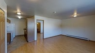 This single-story space presents an open and functional layout designed for comfortable everyday living. The main living area is finished with light hardwood-style flooring that extends throughout, creating a cohesive and easy-to-maintain surface underfoot. Neutral white walls keep the space feeling bright and versatile, providing a blank canvas for personalization.

A partial wall and cabinetry divide the living area from the kitchen zone, which features medium-toned wood cabinetry and tile flooring, offering a practical transition between spaces. The kitchen includes what appears to be standard countertop workspace and storage cabinetry, making efficient use of the available footprint.

A doorway visible from the main living area leads to an additional room, suggesting a separate bedroom or utility space beyond. A baseboard heating unit along the far wall provides climate control, a common and reliable feature in residential spaces across many U.S. regions.

The layout is straightforward and uncluttered, with standard ceiling heights and even lighting throughout. Overall, the space offers a clean, move-in-ready environment with practical finishes suited for functional, day-to-day use.