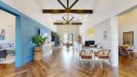 This single-story community clubhouse is designed with a cohesive coastal-modern farmhouse aesthetic, combining shiplap wall paneling, bold blue accent walls, herringbone-patterned wood flooring, and soaring vaulted ceilings throughout the main gathering areas. The entry lobby serves as the central welcome point, featuring a staffed reception desk, rattan seating, an open bookshelf display, and a striking gold geometric chandelier suspended beneath exposed dark wood beams. A yellow neon location-pin logo adds a branded focal point to the shiplap wall. Adjacent to the lobby, a spacious lounge area offers comfortable seating arrangements centered around a wall-mounted TV on a chevron-patterned blue accent wall, creating an inviting space for casual gatherings. A fully equipped coffee and refreshment bar features dark subway tile backsplash, white cabinetry, a dark countertop island with bar seating, open wood shelving, and built-in appliances. A dedicated conference room accommodates a large group around a solid wood farmhouse table, surrounded by leather executive chairs, with ample natural light streaming through floor-to-ceiling sheer-draped windows. The exterior grounds feature a generously sized resort-style pool deck lined with lounge chairs and shade umbrellas, completing this full-featured community amenity space.