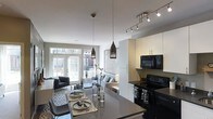 This contemporary single-floor residence showcases a thoughtfully designed open-concept layout that seamlessly connects the kitchen, dining, and living areas into one cohesive living space. The kitchen is outfitted with sleek white cabinetry accented by gold hardware, dark countertops, and black appliances, including a range and over-the-range microwave. A central island with an extended countertop serves as both a food preparation surface and a casual dining spot. Distinctive pendant lights with dark metallic finishes hang above the island, complemented by a modern track lighting system that provides ample task and ambient illumination. The dining area offers a stylish table set for multiple guests, complete with woven placemats and decorative tableware. The living room is anchored by a comfortable sofa and armchair arrangement set atop a patterned area rug, with a decorative plant adding a natural touch. Large French doors flood the space with abundant natural light and offer views of an outdoor area, creating an airy and welcoming ambiance. The neutral color palette of whites, grays, and soft tones throughout gives the space a clean, modern aesthetic.