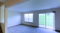 This two-floor residence offers a clean, functional layout designed to maximize comfort and usability. The main living area is a spacious, open room finished with soft wall-to-wall carpeting and neutral-toned walls that create a calm, versatile backdrop for any furnishing style. A large window fitted with horizontal blinds allows abundant natural light to fill the room, while a sliding glass door provides direct access to the outdoors, where a well-maintained green lawn is visible — a welcoming feature that seamlessly connects interior living with outdoor space. The sliding door is framed with a classic grid-style design, adding a subtle architectural detail to the room. The ceiling is smooth and generously proportioned, contributing to the airy feel of the space. Trim and baseboards throughout are finished in a complementary tone, lending a polished, cohesive look. Spread across two floors, the home provides distinct zones for living, offering both shared and private spaces suited to a variety of lifestyles. Overall, the space conveys a bright, approachable character with practical finishes and a strong connection to the natural surroundings outside.