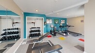 Located in Springdale, AR, this dedicated exercise room offers a thoughtfully designed fitness space optimized for a full-body workout experience. The room features light-toned walls accented by a bold teal feature wall that adds energy and vibrancy to the environment. Large tile flooring with colorful inset squares in red, yellow, and teal provides a dynamic visual element while remaining easy to clean and maintain.

Natural light flows in through multiple windows, including a set of French doors, keeping the space bright and inviting. Recessed and flush-mount ceiling lights ensure excellent illumination throughout. A large wall-mounted mirror spans a significant portion of one wall, enhancing the sense of space and allowing users to monitor their form during workouts.

The room is outfitted with a variety of fitness equipment, including a treadmill, elliptical, recumbent bike, and a multi-use weight bench. A dedicated storage area houses a full rack of kettlebells and additional fitness accessories, keeping the space organized. Framed artwork decorates the teal accent wall, adding a touch of personality. A security camera is also present, contributing to a safe and monitored environment. This exercise room is a complete, functional fitness facility.