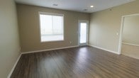 This single-floor space offers a bright and functional layout designed with simplicity and comfort in mind. The main living area is finished with rich, dark hardwood-style flooring that adds warmth and depth to the room, complementing the neutral beige-toned walls throughout. A large window fitted with horizontal blinds allows generous amounts of natural light to fill the space, creating an open and airy atmosphere. A glass-paneled exterior door provides additional light and direct access to the outdoors, further enhancing the sense of openness. The ceiling features recessed lighting and visible air vents, ensuring both practical illumination and effective climate control. Clean white baseboards and trim provide a crisp contrast to the warm flooring and walls, lending a polished, well-maintained appearance. The layout is open and unobstructed, offering flexible potential for a variety of furnishing arrangements and uses. Overall, the space presents a modern yet understated aesthetic, combining functional design elements with a welcoming, neutral palette that is well-suited for a range of purposes.