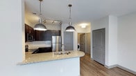 This single-floor, 3-bedroom, 2-bathroom residence showcases a well-appointed open-concept layout that centers around a striking kitchen. The kitchen serves as the heart of the space, featuring a large curved island topped with speckled granite-style countertops and an undermount double sink. Dark espresso cabinetry lines the walls, providing ample storage and creating a bold contrast against the light-colored counters and white subway tile backsplash. Stainless steel appliances, including a refrigerator and built-in microwave, add a sleek, modern touch.

Overhead, industrial-style pendant lights hang above the island, complemented by a track lighting system that ensures the workspace is well-lit. The kitchen flows seamlessly into the surrounding living areas, enhanced by warm-toned hardwood-style flooring that extends throughout the main space.

A hallway visible from the kitchen leads to the three bedrooms and two bathrooms, with in-unit laundry conveniently tucked nearby. Gray interior doors with detailed paneling add an elegant, cohesive design element throughout. The overall ambiance is contemporary and functional, balancing stylish aesthetics with practical, everyday livability.
