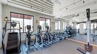 This fitness center offers a bright and well-appointed workout environment designed to support a range of cardio-focused exercise routines. The space is characterized by its impressively high ceilings, which feature exposed architectural ribbing and pendant track lighting that adds both functionality and visual interest overhead. Large, black-framed windows line the perimeter walls, allowing abundant natural light to fill the room while providing views of the surrounding exterior. Wall-mounted lantern-style sconces complement the pendant lighting, creating a warm and inviting atmosphere despite the facility's modern aesthetic. A full-length mirror spans one wall, effectively doubling the perceived depth of the space and enhancing the sense of openness. The flooring consists of a combination of protective rubber matting beneath the equipment and smooth hard flooring in the surrounding areas. The cardio equipment lineup includes multiple elliptical trainers and rowing machines, all arranged in organized rows for ease of use. The overall ambiance is clean, airy, and energizing, making it a well-designed space for residents or members seeking a convenient and comfortable fitness facility.