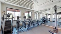 This fitness center offers a bright and well-appointed workout environment designed to support a range of cardio-focused exercise routines. The space is characterized by its impressively high ceilings, which feature exposed architectural ribbing and pendant track lighting that adds both functionality and visual interest overhead. Large, black-framed windows line the perimeter walls, allowing abundant natural light to fill the room while providing views of the surrounding exterior. Wall-mounted lantern-style sconces complement the pendant lighting, creating a warm and inviting atmosphere despite the facility's modern aesthetic. A full-length mirror spans one wall, effectively doubling the perceived depth of the space and enhancing the sense of openness. The flooring consists of a combination of protective rubber matting beneath the equipment and smooth hard flooring in the surrounding areas. The cardio equipment lineup includes multiple elliptical trainers and rowing machines, all arranged in organized rows for ease of use. The overall ambiance is clean, airy, and energizing, making it a well-designed space for residents or members seeking a convenient and comfortable fitness facility.