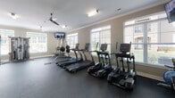 Located at 159 W Kinderton Way in Bermuda Run, NC, this well-appointed fitness center — branded as Commit Bermuda Run — offers a comprehensive, single-floor workout facility designed to support a full range of exercise routines. The space is bathed in natural light, courtesy of large windows with white blinds that line multiple walls, creating a bright and energizing atmosphere throughout.

The main cardio area is generously sized, housing a row of True treadmills and elliptical machines arranged to maximize floor space, while ceiling fans overhead ensure comfortable airflow during workouts. A wall-mounted flat-screen TV is positioned for easy viewing from the cardio equipment.

A dedicated free weights section features a multi-tiered dumbbell rack stocked with a wide range of weights, accompanied by an adjustable workout bench and a vertical knee raise/dip station for strength training versatility. The flooring throughout is durable rubber gym matting, providing both safety and comfort.

A transitional area near the entry includes a mounted SmartMat exercise guide, a seating bench, and dual water fountains for convenient hydration. Artwork on the wall adds a welcoming touch. Crown molding and neutral taupe walls lend the space a polished, finished feel that elevates it beyond a standard fitness room. The facility is well-lit with both natural and track lighting throughout.