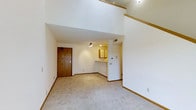 This two-story space makes an immediate impression with its dramatically tall ceilings in the main living area, creating an expansive, airy atmosphere. The open-concept main floor features plush, light-toned carpeting throughout, contributing to a neutral and welcoming aesthetic. White walls and warm wood-toned trim work together to provide a clean, classic finish across the space.

The living area flows naturally toward a pass-through kitchen counter, which offers both a visual connection between rooms and a practical serving surface. The kitchen features wood-toned cabinetry and is efficiently laid out, providing functional storage and workspace. A ceiling-mounted light fixture illuminates the kitchen area, while the open sightlines to the living space keep the overall feel bright and connected.

A solid wood entry door adds a warm, grounded contrast to the otherwise light interior palette. An upper-level overlook — visible from the main floor — reinforces the vertical openness of the space and hints at additional functional square footage above. The second floor is accessible via a staircase, completing this multi-level layout designed to maximize both volume and livability.