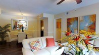 This single-floor residence offers a warm and inviting atmosphere defined by its cheerful color palette and thoughtfully curated interiors. The living area serves as the heart of the space, centered around a plush white sofa accented with colorful decorative pillows in coral and Southwestern-patterned prints. A ceiling fan overhead provides both function and a casual, comfortable feel, while the textured ceiling adds subtle architectural character.

The walls are painted in soft, warm tones that complement the vibrant large-scale abstract artwork displayed prominently, adding an artistic focal point to the room. Fresh floral arrangements featuring birds of paradise and yellow blooms bring natural color and life to the space.

An open sightline connects the living area to a compact dining nook and the kitchen beyond, where cabinetry and appliances are partially visible, suggesting a practical and efficient layout. A hallway extends further into the home, hinting at additional private rooms. Neutral base tones throughout allow the colorful accents — from artwork to flowers to textiles — to stand out, creating a space that feels lively yet cohesive.