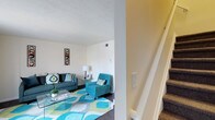 This two-floor residence offers a well-organized layout that balances functionality with a cheerful, modern aesthetic. The main level features an inviting living area bathed in natural light, showcasing light-toned walls that create an open and airy atmosphere. The living space is thoughtfully arranged with ample room for seating and relaxation, anchored by a bold, multicolored geometric area rug in shades of teal, green, and gray that adds a vibrant focal point to the room. A glass-topped coffee table and a combination of a sofa and accent chair in coordinating teal tones contribute to the cohesive, contemporary style. Decorative wall art and a classic table lamp add character and warmth to the space. A carpeted staircase, visible from the entry point, provides a smooth transition to the upper level, featuring clean white railings that complement the neutral color palette throughout. The textured ceiling adds a subtle architectural detail, while the dark hardwood-style flooring on the main level grounds the space with a touch of elegance. Overall, this home presents a bright, well-appointed interior designed to accommodate both relaxed living and everyday functionality across its two floors.