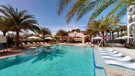 This single-level amenity space centers around a generously sized resort-style swimming pool with crystal-clear turquoise water, creating an inviting and vacation-like atmosphere. The pool area is thoughtfully designed with a smooth surrounding deck that accommodates multiple lounge chairs and shaded umbrella stations, offering ample space for both sunbathing and relaxed gatherings. Tall, swaying palm trees line the perimeter, enhancing the tropical ambiance and providing natural framing against a bright blue sky. At the far end of the pool, a distinctive white stucco structure with elegant arched openings serves as an architectural focal point, lending a Mediterranean or Spanish Colonial character to the space. A water feature element is also visible near the structure, adding a dynamic and soothing quality to the environment. The combination of lush greenery, warm natural light, and the shimmering pool surface creates a serene, resort-caliber outdoor experience. Overall, the space exudes a relaxed yet polished aesthetic, making it well-suited for leisure, socializing, and outdoor recreation in a richly landscaped setting.