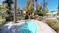 This inviting outdoor amenity space embodies classic Spanish Colonial architectural design, characterized by smooth stucco walls finished in warm cream tones, decorative arched colonnades, and red-tiled rooflines visible on the surrounding multi-story buildings. The centerpiece of the space is a generously sized swimming pool with a distinctive curved entry wading area, finished in clear turquoise water and surrounded by a broad, light-toned stone pool deck ideal for lounging. A row of poolside chaise lounges lines the deck beneath shaded green umbrella cabanas, offering comfortable outdoor seating in the sun-drenched environment. The pool area is accessed through an elegant arched entryway with decorative wrought-iron gating, adding a refined, resort-like sense of arrival. Towering date palms and mature shade trees create a lush, tropical canopy overhead, lending the space a private, oasis-like ambiance. Curved walkways wind through the manicured landscaping, with young palms and desert-adapted plantings adding texture and greenery throughout. The surrounding buildings feature private balconies with iron railings that overlook the pool courtyard, reinforcing a cohesive and immersive resort atmosphere. Under abundant natural sunlight and clear blue skies, this outdoor space delivers a tranquil and visually striking retreat.