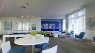 This contemporary single-floor space is designed with a clean, modern aesthetic that immediately captures attention. The focal point of the room is a striking bold blue tiled accent wall, which serves as a dynamic backdrop for a row of integrated flat-screen displays, adding both a visual statement and a functional media element to the environment. The open-plan layout is thoughtfully arranged with distinct zones for different uses. A casual dining area in the foreground features sleek round white tables paired with modern chairs accented in deep blue cushions, creating a cohesive color palette throughout. The lounge area beyond is furnished with neutral-toned sofas and ottomans, offering comfortable and inviting seating. Tall, floor-to-ceiling windows dressed with sheer white curtains line the perimeter, allowing an abundance of natural light to permeate the interior and providing views of the surrounding urban landscape. Wood-plank flooring in a cool gray tone grounds the space with warmth and texture, while recessed and pendant lighting enhances the bright, airy atmosphere. Touches of greenery, including potted plants on the tables, bring a fresh, natural element to the polished interior. The overall ambiance is sophisticated, welcoming, and well-suited for social gathering or relaxation.