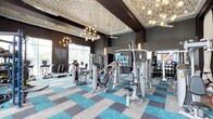 This contemporary fitness center is thoughtfully designed to deliver both functionality and style in equal measure. The space makes an immediate impression with its elevated tray ceilings adorned with an intricate geometric textured finish, complemented by a dramatic combination of globe cluster chandeliers and a sleek ceiling fan that together create a bright, motivating atmosphere. Dark accent walls provide a bold contrast to the lighter ceiling elements, lending the space a refined, upscale character. The flooring features a striking patchwork of teal and gray carpet tiles, adding a vibrant splash of color that energizes the environment. Large windows along the perimeter invite natural light and offer views to the outdoors, enhancing the sense of openness. The fitness floor is comprehensively outfitted with a full array of professional-grade equipment, including treadmills, ellipticals, stationary bikes, multi-station cable machines, and a dedicated free weights area with an extensive dumbbell rack. A wall-mounted flat-screen television is centrally positioned, keeping members entertained during their workouts. Mirrored walls expand the visual space and support proper form during strength training. Overall, this fitness center strikes an impressive balance between high-end design and practical, well-organized workout amenities.