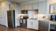 This thoughtfully designed single-floor studio offers a seamless open layout that integrates the living, sleeping, and kitchen areas into one cohesive space. The kitchen is a standout feature, showcasing soft gray shaker-style cabinetry, granite countertops, and a striking hexagonal tile backsplash. A full suite of stainless steel appliances — including a refrigerator, electric range, over-the-range microwave, and dishwasher — provides a fully functional cooking environment. Under-cabinet lighting adds a polished finishing touch. A built-in desk with matching granite countertop offers a dedicated workspace seamlessly integrated into the kitchen area. Rich hardwood-style flooring flows throughout, grounding the neutral, warm color palette. The sleeping area includes a spacious closet with built-in shelving, while a wall-mounted television and comfortable seating create a relaxed living zone. Large windows invite natural light and provide pleasant outdoor views, enhancing the open ambiance. The bathroom features a granite-topped vanity with white cabinetry, an undermount sink, and a walk-in shower, completing this well-finished and functional space.
