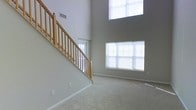 This two-story home makes an immediate impression with its soaring double-height ceiling in the main living area, creating a grand sense of openness and volume. The space is anchored by a wood-railed staircase with natural oak balusters and a turned newel post, providing both functional access to the upper level and a classic architectural focal point. Neutral gray walls and light-toned carpeting run throughout, contributing to a clean, cohesive aesthetic that complements a variety of furnishing styles. Natural light is a defining feature of this living space, delivered through a large multi-pane window on the main level and a tall clerestory window positioned high on the upper wall, flooding the interior with daylight at multiple heights. White window blinds provide light control while maintaining the bright, airy atmosphere. Ventilation infrastructure is integrated into the upper wall, and standard electrical outlets are distributed throughout the room. The overall ambiance is open, neutral, and well-lit, offering a versatile canvas suited to a range of interior design approaches.