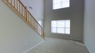 This two-story home makes an immediate impression with its soaring double-height ceiling in the main living area, creating a grand sense of openness and volume. The space is anchored by a wood-railed staircase with natural oak balusters and a turned newel post, providing both functional access to the upper level and a classic architectural focal point. Neutral gray walls and light-toned carpeting run throughout, contributing to a clean, cohesive aesthetic that complements a variety of furnishing styles. Natural light is a defining feature of this living space, delivered through a large multi-pane window on the main level and a tall clerestory window positioned high on the upper wall, flooding the interior with daylight at multiple heights. White window blinds provide light control while maintaining the bright, airy atmosphere. Ventilation infrastructure is integrated into the upper wall, and standard electrical outlets are distributed throughout the room. The overall ambiance is open, neutral, and well-lit, offering a versatile canvas suited to a range of interior design approaches.