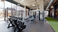 Located at 367 Orange St in New Haven, CT, this single-floor fitness center combines industrial design with a comprehensive, well-equipped workout environment. The space is defined by its exposed concrete columns and ceilings, visible ductwork, and large floor-to-ceiling windows that flood the interior with natural light and offer views of the surrounding New Haven streetscape.

The cardio zone features a full row of treadmills and stationary cycling bikes positioned along the expansive window wall, allowing members to exercise with an abundance of daylight. Adjacent to this area, the free weights section is stocked with a wide range of dumbbells, barbells, and weight plates, supported by power racks and adjustable benches suited for a variety of strength training exercises.

A dedicated functional training zone features an artificial turf runway alongside wall-mounted shelving stocked with medicine balls and agility equipment, ideal for sport-specific and dynamic workouts. Cable machines and multi-functional training stations are distributed throughout the floor, providing versatile resistance training options.

The rubber flooring throughout ensures safety and durability, while pendant lighting supplements the natural light to maintain a bright, energizing atmosphere. Full-length mirrors are strategically placed to enhance the sense of space and support proper form. The overall ambiance is modern, motivating, and purpose-built for serious fitness.