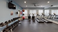 This fitness center is a comprehensive, single-floor workout facility designed to accommodate a wide range of exercise needs. The space is finished with durable rubber flooring throughout, providing a safe and practical surface for all types of training. The walls are painted in a cool blue tone, contributing to an energizing yet focused atmosphere, while a textured ceiling with recessed lighting and ceiling fans ensures the space remains well-lit and well-ventilated.

The cardio zone features multiple treadmills and elliptical machines positioned along one wall, benefiting from natural light streaming through several windows fitted with horizontal blinds. Adjacent to the cardio equipment, a well-stocked dumbbell rack offers a broad range of free weights for strength training, accompanied by stability and medicine balls for functional fitness exercises. A wall-mounted flat-screen television provides entertainment during workouts.

The strength training area is generously outfitted with multiple cable and selectorized weight machines, along with adjustable benches and TRX suspension training equipment anchored to the wall. Full-length mirrors line one side of the room, amplifying the sense of space and allowing users to monitor their form. An exterior door with glass panels provides direct access to the outdoors, adding convenience and a connection to the surrounding environment.