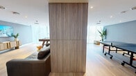 Located at 16395 Biscayne Boulevard in North Miami Beach, Florida, this amenity lounge is a well-appointed, single-floor social space designed for both relaxation and recreation. The room is finished with light-toned hardwood flooring throughout, contributing to a warm and cohesive aesthetic that pairs seamlessly with the modern interior design.

The space is thoughtfully divided into distinct functional zones. A lounge area features a dark leather sofa, contemporary curved accent chairs, and a pair of wall-mounted flat-screen televisions set atop sculptural wooden media consoles, creating an inviting entertainment hub. Abstract artwork adorns the gray accent walls, adding a curated, gallery-like quality to the atmosphere.

A central wood-paneled architectural column serves as a visual divider, separating the lounge from the recreation zone, which houses both a pool/air hockey table and a ping-pong table. Floor-to-ceiling sheer curtains line the perimeter windows, filtering abundant natural light while maintaining a bright, airy feel. Recessed ceiling lighting supplements the natural illumination throughout.

The entry area features glass double doors, additional seating, and greenery that enhances the welcoming atmosphere. The overall design strikes a balance between contemporary sophistication and casual comfort, making it a versatile communal space suited for social gatherings and leisure activities alike.