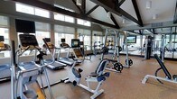 This single-floor fitness center is thoughtfully designed to offer a comprehensive workout experience in a bright, welcoming environment. The space is anchored by a dramatic vaulted ceiling with bold, dark exposed beam trusses that lend an architectural character and a sense of generous vertical volume to the room. A continuous row of large windows lines the upper walls, flooding the interior with abundant natural light and offering views of the surrounding exterior, creating an open and energizing atmosphere.

The flooring throughout is a durable, sport-appropriate surface well-suited for high-traffic fitness use. The cardio section is well-appointed with multiple treadmills featuring integrated digital displays, upright and recumbent stationary bikes, and additional aerobic equipment. The strength training area includes a multi-station cable and weight machine system, free weights, and a rack with weight plates, providing a full range of resistance training options. Large flat-screen monitors are mounted within the space, likely for entertainment or instructional purposes during workouts.

The layout is spacious and uncluttered, allowing for comfortable movement between stations. The combination of high ceilings, natural light, modern equipment, and thoughtful design makes this fitness center a functional and motivating space for users of all fitness levels.