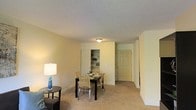 This single-floor residence offers a practical and comfortable layout designed to make the most of its living areas. The combined living and dining space serves as the heart of the home, featuring wall-to-wall carpeting that adds warmth underfoot and a neutral color palette that lends a clean, inviting ambiance throughout.

The dining area comfortably accommodates a four-seat dining set, positioned beneath flush-mounted ceiling lighting that provides functional illumination. A built-in shelving unit is recessed into the wall nearby, offering convenient display and storage space while adding a subtle architectural detail to the room. The textured ceiling adds a classic residential character to the space.

Adjacent to the dining area, the living space provides room for seating and relaxation, with a side table and lamp contributing to a cozy atmosphere. A entry door is visible along the far wall, offering direct access and contributing to the flow of the layout. The neutral, light-toned walls throughout the space enhance the sense of openness, while the consistent flooring and trim work unify the design. Overall, this residence presents a straightforward, well-maintained interior with a focus on everyday functionality and comfort.