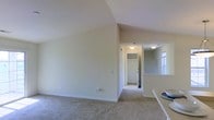This single-floor residential space offers a clean, neutral aesthetic throughout, with soft beige walls and wall-to-wall carpeting that create a warm and cohesive atmosphere. The main living area is distinguished by its vaulted ceiling, which angles upward to add a sense of openness and volume to the room. The elevated ceiling height contributes to an airy feel, making the space appear larger and more expansive than a standard flat-ceiling layout.

A hallway extends from the main living area, connecting to additional rooms and providing a practical flow through the home. Visible from the main space, a reach-in closet with a rod and built-in shelving offers dedicated storage. Flush-mount ceiling light fixtures illuminate the hallway and adjoining areas, providing consistent and even lighting throughout.

To one side, a glimpse of a window allows natural light to filter into an adjacent area, brightening the interior. A partial white countertop or ledge is also visible, hinting at a kitchen or bar area nearby. The overall palette of whites and soft neutrals throughout the walls, trim, and ceilings gives the space a clean, move-in-ready appearance that is both functional and welcoming.