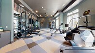 Located in Norcross, Georgia, this fitness center offers a thoughtfully designed workout space across a single floor. The room is finished with a distinctive large-scale checkered carpet in neutral tones of beige, gray, and brown, providing a visually striking foundation that ties the space together. Walls painted in muted teal and gray tones lend a calm, modern aesthetic, complemented by decorative vertical shiplap paneling and arched wall niches adorned with framed artwork, adding character beyond a typical gym environment. The tray ceiling, finished in dark gray with recessed lighting and a ceiling fan, creates a sense of architectural depth while ensuring the space remains bright and well-ventilated. A full-length mirror spans one wall, effectively doubling the visual footprint of the room and enhancing the sense of openness. Natural light filters in through large windows, offering views of the surrounding landscape. Equipment includes a row of treadmills, an elliptical, a rowing machine, a multi-station cable/strength machine, a power rack with bench, a dumbbell rack, and additional cardio machines. A wall-mounted television is also present. The overall ambiance is polished and inviting, balancing functionality with interior design sensibility.