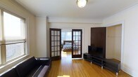 This well-appointed one-bedroom, one-bathroom residence offers a thoughtfully organized single-floor layout. The living area features warm hardwood flooring that extends throughout the space, creating a cohesive and inviting atmosphere. Crown molding adds a classic architectural touch, lending the unit a sense of timeless character. A standout feature is the pair of elegant French doors with dark wood frames and divided glass panes that connect the living room to the bedroom, allowing natural light to flow freely between the two spaces while offering the option for privacy. The bedroom benefits from large windows that bring in ample natural light and offer pleasant outdoor views. The living area provides comfortable room for seating and entertainment, with space for a sofa and media console. The dark wood accents of the French doors and built-in elements contrast attractively with the warm tones of the hardwood floors and the soft neutral walls. Overall, the residence combines classic detailing with a practical layout, creating a bright and functional living environment suitable for everyday comfort.