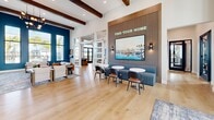 The Yardly Ellenton leasing and amenity center at 4180 Yardly Dr in Palmetto, Florida is a thoughtfully designed single-story space that blends contemporary style with a welcoming, community-focused atmosphere. Soaring ceilings accented by exposed dark wood beams lend an open, airy feel throughout, while light hardwood flooring ties the layout together seamlessly.

The reception area greets visitors with a striking living moss wall display, a beverage station, and a staffed welcome desk framed by sleek glass-paneled doors bearing the Yardly Ellenton by Taylor Morrison branding. Adjacent to the entry, a lounge area features a dramatic floor-to-ceiling marble tile fireplace wall with a mounted flat-screen TV, a cozy seating arrangement, and oversized windows overlooking outdoor pool views — bathing the room in natural light.

A social lounge with a "Find Your Home" feature wall showcases slatted wood paneling, coastal artwork, and banquette seating paired with bistro tables. The open kitchen area includes a waterfall-edge island with bar seating, pendant lighting, and a bold Tampa, Florida mural that adds a vibrant local character. Private booth seating nooks with blue upholstery, greenery, and large windows offer intimate spaces for one-on-one meetings or quiet work. Throughout, the design balances energetic branding with comfortable, functional spaces.