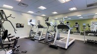 This single-floor fitness center is designed to support a well-rounded workout experience. The space features a clean, functional layout with light-colored walls accented by a green feature wall, creating an energizing atmosphere. Overhead, a drop ceiling with recessed fluorescent panel lighting ensures the room is evenly and brightly illuminated throughout.

The cardio area is outfitted with a row of Precor machines, including treadmills and elliptical cross-trainers, each equipped with individual media screens for user convenience. The machines are arranged on durable dark rubber flooring, which provides both traction and protection suited to high-impact use.

A large wall-to-wall mirror runs along one side of the room, visually expanding the space and allowing users to monitor their form during workouts. Natural light filters in through a window on the adjacent wall, adding a welcoming brightness to the environment.

The overall ambiance is clean, modern, and well-maintained, making it a practical and inviting space for residents or guests seeking convenient access to fitness facilities.