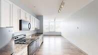 Located at 3101 Euclid Ave in Cleveland's vibrant midtown corridor, this efficiently designed single-floor unit offers a seamless open-plan layout combining the kitchen and living area into one cohesive space. The kitchen is well-appointed with crisp white shaker-style cabinetry extending to the ceiling, granite countertops, and a full suite of black appliances including a refrigerator, electric range, built-in microwave, and dishwasher. A stainless steel sink with a modern faucet completes the functional workspace. Track lighting overhead adds a contemporary touch while providing excellent task illumination throughout the kitchen and living zone. Light-toned wood-style flooring runs continuously throughout the unit, enhancing the sense of openness and flow. A hallway connects the main living area to additional rooms, lined with recessed lighting and clean white walls. The living area benefits from a large picture window fitted with horizontal blinds, framing urban views of the Cleveland cityscape and filling the space with natural light. The overall aesthetic is clean and modern, with soft gray walls and minimalist finishes creating a bright, neutral palette throughout.