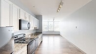 Located at 3101 Euclid Ave in Cleveland's vibrant midtown corridor, this efficiently designed single-floor unit offers a seamless open-plan layout combining the kitchen and living area into one cohesive space. The kitchen is well-appointed with crisp white shaker-style cabinetry extending to the ceiling, granite countertops, and a full suite of black appliances including a refrigerator, electric range, built-in microwave, and dishwasher. A stainless steel sink with a modern faucet completes the functional workspace. Track lighting overhead adds a contemporary touch while providing excellent task illumination throughout the kitchen and living zone. Light-toned wood-style flooring runs continuously throughout the unit, enhancing the sense of openness and flow. A hallway connects the main living area to additional rooms, lined with recessed lighting and clean white walls. The living area benefits from a large picture window fitted with horizontal blinds, framing urban views of the Cleveland cityscape and filling the space with natural light. The overall aesthetic is clean and modern, with soft gray walls and minimalist finishes creating a bright, neutral palette throughout.