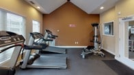 Located at 4085 Warner Ave in Landover Hills, Maryland, this single-floor fitness center is thoughtfully designed to support a well-rounded workout experience. The space is defined by its striking vaulted ceiling, which rises to a dramatic peak and is accented by a large A-frame window that floods the room with natural light and offers pleasant views of surrounding greenery. Recessed lighting throughout ensures the space remains bright and well-lit at all hours.

The facility is equipped with a comprehensive selection of cardio machines, including multiple treadmills, elliptical trainers, and a recumbent stationary bike — all arranged to maximize floor space and ease of movement. A multi-function cable strength machine provides options for resistance and functional training, rounding out the equipment offerings. The flooring is a durable dark gym-grade material suited for high-traffic exercise use.

Warm earth-toned walls in shades of golden yellow and terracotta create an energizing yet comfortable ambiance. Multiple windows fitted with horizontal blinds allow for adjustable light control throughout the day. A glass-paned entry door connects the fitness area to an adjacent lounge or common area, adding to the overall sense of openness. The layout is efficient and welcoming, making it a practical and inviting space for daily fitness routines.