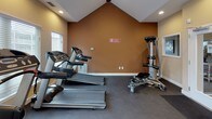 Located at 4085 Warner Ave in Landover Hills, Maryland, this single-floor fitness center is thoughtfully designed to support a well-rounded workout experience. The space is defined by its striking vaulted ceiling, which rises to a dramatic peak and is accented by a large A-frame window that floods the room with natural light and offers pleasant views of surrounding greenery. Recessed lighting throughout ensures the space remains bright and well-lit at all hours.

The facility is equipped with a comprehensive selection of cardio machines, including multiple treadmills, elliptical trainers, and a recumbent stationary bike — all arranged to maximize floor space and ease of movement. A multi-function cable strength machine provides options for resistance and functional training, rounding out the equipment offerings. The flooring is a durable dark gym-grade material suited for high-traffic exercise use.

Warm earth-toned walls in shades of golden yellow and terracotta create an energizing yet comfortable ambiance. Multiple windows fitted with horizontal blinds allow for adjustable light control throughout the day. A glass-paned entry door connects the fitness area to an adjacent lounge or common area, adding to the overall sense of openness. The layout is efficient and welcoming, making it a practical and inviting space for daily fitness routines.