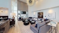 This elegantly designed social clubroom offers a refined communal space defined by dramatic double-height ceilings and expansive floor-to-ceiling windows draped in sheer white curtains that flood the interior with natural light and frame sweeping outdoor views. The light-toned hardwood flooring runs throughout, anchoring a cohesive, contemporary aesthetic.

The main lounge area is centered around generous sectional sofas in soft gray, paired with sculptural black coffee tables beneath a striking sputnik chandelier that serves as a bold focal point. A secondary seating arrangement features deep navy velvet club chairs surrounding a marble-top round table, accompanied by a large organic stone sculpture adding an artistic touch.

A dedicated kitchenette and bar area includes sleek gray cabinetry, a pearl mosaic tile backsplash, dual wall-mounted flat-screen televisions, a microwave, and a large white quartz island with pendant lighting — ideal for entertaining. A cozy dining nook accommodates intimate gatherings with a marble oval table, plush banquette seating, cowhide accent chairs, and its own sputnik chandelier overhead.

Throughout the space, curated artwork and thoughtfully selected furnishings contribute to a luxurious yet welcoming atmosphere, making this clubroom a standout amenity for social interaction and relaxation.