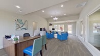 This single-story space functions as a welcoming community or leasing office, thoughtfully designed to balance professional functionality with a comfortable, inviting atmosphere. The open-concept layout is unified by light gray hardwood-style flooring and neutral wall tones, creating a clean, contemporary aesthetic throughout.

The reception area features a warm wood-toned desk with seating for guests, positioned near large windows that draw in generous natural light and offer views of the surrounding landscaping. A decorative orb chandelier anchors the central lounge area, where vivid blue upholstered armchairs with green-striped accent pillows are arranged around a round wood-and-white coffee table, encouraging a relaxed, welcoming environment.

Toward the back, a workstation with a desktop computer sits beneath a wall-mounted flat-screen television, accompanied by a beverage station with a coffee maker and compact wine refrigerator set into white cabinetry with granite-style countertops. Glass interior windows allow visibility into private office or conference areas without compromising separation.

Vaulted ceilings in the main gathering space enhance the sense of openness, while recessed lighting and large exterior-facing doors and windows ensure the interior remains bright and connected to the outdoors.