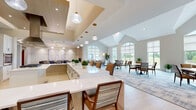 This generously sized community room in Menomonee Falls, Wisconsin, offers a thoughtfully designed, single-floor layout that seamlessly blends dining, living, and kitchen functions. The main gathering area is anchored by soaring vaulted ceilings and a striking arched window that floods the space with natural light, offering views of the surrounding landscape. Patterned area rugs layer warmth over light-toned flooring, while an assortment of comfortable seating arrangements invites conversation and relaxation. A living area features a tile-surround fireplace, a mounted flat-screen television, and refined furnishings beneath dramatic floor-to-ceiling windows. The dining room is distinguished by a linear pendant light fixture, a solid wood table, upholstered chairs, and an elegant tile fireplace wall accented with curated artwork. The kitchen showcases white shaker cabinetry, quartz countertops, an iridescent tile backsplash, open wood shelving, stainless steel appliances, and a large island with bar seating beneath globe pendant lights. Additional smaller seating areas with carpeted floors and large windows provide intimate spaces for smaller gatherings. Throughout, the neutral warm palette, quality materials, and abundant natural light create a welcoming and sophisticated communal environment.