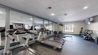 This single-floor fitness center is designed to support a comprehensive workout experience in a clean, functional environment. The main exercise area is outfitted with multiple treadmills, a rowing machine, and a multi-station cable strength machine, all arranged on durable rubber flooring suited for high-traffic use. A full wall of floor-to-ceiling mirrors lines one side of the cardio and strength zone, visually expanding the space while allowing users to monitor their form during exercise. A dumbbell rack stocked with free weights and medicine balls rounds out the strength training offerings. Recessed lighting and a mounted flat-screen television add comfort and convenience to the workout environment. Adjacent to the main gym area is a bright, open flex room with large multi-pane windows on two walls that invite generous natural light and outdoor views of surrounding greenery. This versatile space features the same durable flooring and offers ample room for stretching, yoga, or group fitness activities. A wall-mounted folded mat provides additional functional utility. The facility is accessed through a glass-paneled entryway with gray-framed doors, maintaining a cohesive, modern aesthetic throughout. The overall design is practical and well-appointed, providing everything needed for a thorough fitness routine.