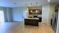 This single-floor space showcases an open-concept layout that seamlessly connects the kitchen and living areas, creating a bright and functional environment. The kitchen is a standout feature, anchored by a large central island with a granite countertop and an integrated sink, providing ample workspace and serving as a natural focal point. Dark espresso-toned cabinetry contrasts elegantly with the light granite surfaces, while stainless steel appliances, including a range and built-in microwave, add a contemporary edge. Three pendant lights hang above the island, complementing the recessed lighting throughout to create a warm, well-lit atmosphere. Light-toned hardwood flooring extends across the open living area, enhancing the sense of spaciousness and contributing to the bright, airy feel of the space. A doorway visible from the main area leads into an adjoining carpeted room, offering a natural separation of living zones. Neutral wall tones throughout provide a clean, versatile backdrop. The overall design balances modern aesthetics with practical functionality, making it a well-appointed and welcoming living space.