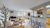 This single-floor residence is thoughtfully laid out to maximize functionality and comfort. Light hardwood flooring runs throughout the main living areas, creating a cohesive and warm aesthetic. The open-concept living and dining area is bright and welcoming, with multiple windows that invite natural light and offer views of surrounding greenery. A wall-mounted air conditioning unit provides climate control, and baseboard heating is present throughout. The living area flows seamlessly into a dining nook, complete with a glass-top table and modern seating, accented by a round wall mirror and decorative greenery.

The kitchen is semi-open to the living space, separated by a dark granite breakfast bar with bar seating. It features light wood cabinetry, dark granite countertops, a stainless steel sink, and full appliances including a refrigerator, stove, and microwave, all illuminated by track lighting overhead.

The bedroom is generously sized, featuring hardwood floors, a window with natural light, and direct access to a spacious walk-in closet outfitted with built-in shelving. The bathroom is finished with tile flooring, a dark vanity with a light countertop, and clean, neutral tones throughout.
